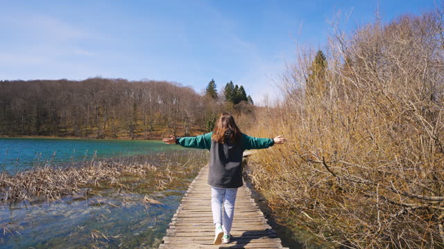 SLO MO Rear View of Carefree Female Tourist with Arms Outstretched Walking on Boardwalk at Plitvice Lakes National Park on Sunny Day