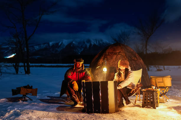 A mixed race couple relax by their tent and camp fire at night with the ground covered in snow.
