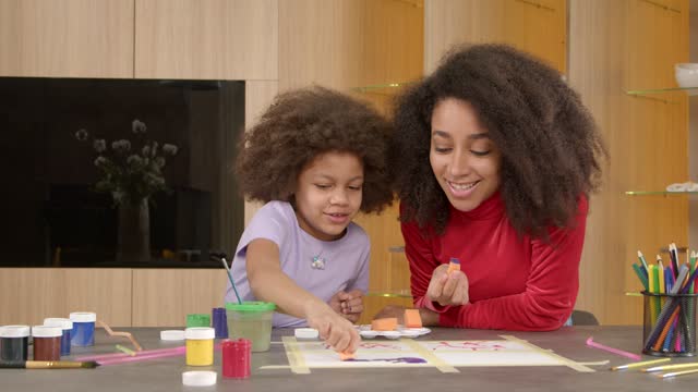 Caring pretty black mother teaching little daughter to sponge painting indoor