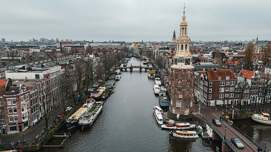 Aerial view of Montelbaanstoren Clock Tower in Amsterdam, Montelbaanstoren tower in Amsterdam, aerial city view of Montelbaanstoren Tower, aerial view of narrow canals of Amsterdam, popular place in Amsterdam\n\nThe Montelbaanstoren is a tower on bank of the Oudeschans – a canal in Amsterdam. The original tower was built in 1516 as part of the Walls of Amsterdam for the purpose of defending the city and the harbour. The top half, designed by Hendrick de Keyser, was extended to its current, decorative form in 1606. Since then the tower has been 48m tall.\n\nBecause the 3rd Duke of Alba proposed incorporating the tower into a castle (the Monte Albano) the tower became known in Dutch as the Monte Albano Toren. Over the years this became garbled to \