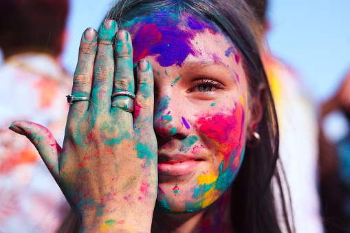 Close-up portrait of a joyful blonde girl with hands and face covered in paint at the Holi festival, celebrating the holiday.
