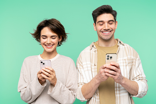 Portrait photo of smiling, attractive pair, using smart phones for online communication, typing, and posing isolated on a turquoise background, focused on their screens