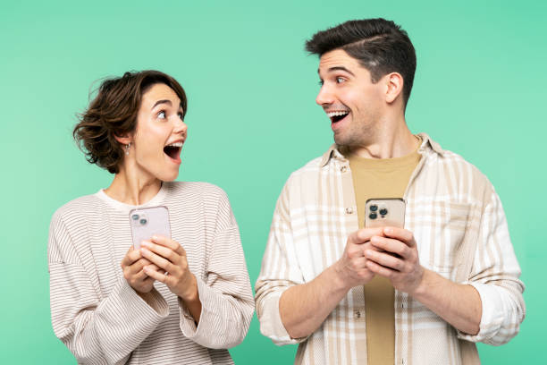 Smiling shocked people holding mobile phones, looking each other, and screaming Portrait photo of smiling, shocked people holding mobile phones, looking each other, and screaming, isolated on a teal background. Online shopping concept surprised men stock pictures, royalty-free photos & images
