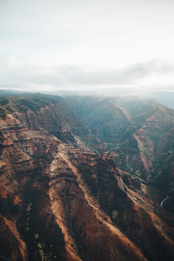 Embark on a journey into the heart of Waimea Canyon, Kauai, Hawaii, USA, with this breathtaking aerial view capturing the rugged beauty of the landscape on a cloudy day. Despite the overcast skies, rays of sunlight break through the clouds, casting dramatic light into the deep ravines of the canyon. The iconic red earth, characteristic of the island, shines vibrantly amidst the lush greenery, showcasing the natural beauty sculpted by the forces of nature. This awe-inspiring image serves as a testament to the remarkable power of nature in shaping the island's breathtaking terrain.