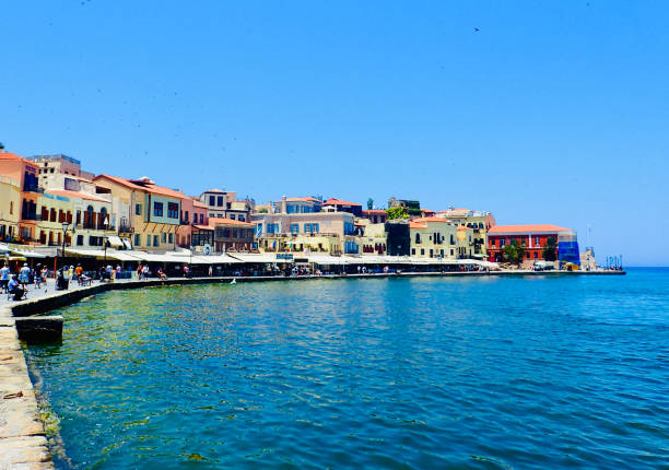 The seaside promenade of Chania bustles with life, showcasing a picturesque array of waterfront cafes and traditional Greek buildings under a clear blue sky.