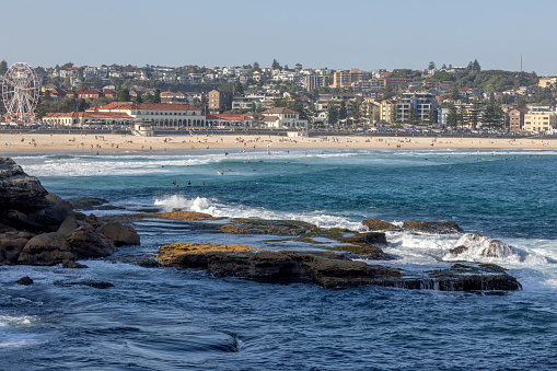 Waves washing up on rocks in the foreground, before iconic Bondi Beach and Bondi Pavilion, in Sydney, Australia. People enjoying the sand and water.