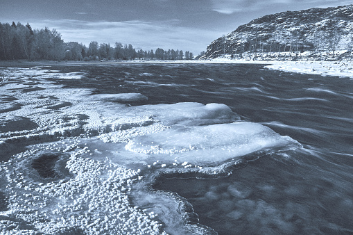 A river flows with ice along its shore, framed by a majestic mountain in the background. The sky is filled with clouds, creating a beautiful natural landscape. Russia, Chuisky ridge, Katun river.