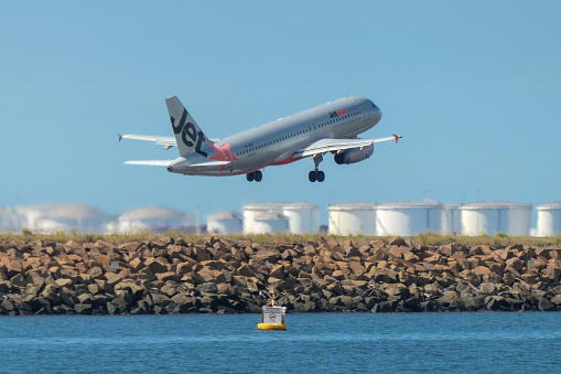 A Jetstar Airbus A320-232 plane, registration VH-VQH, taking off from Sydney Kingsford-Smith Airport and heading to Hobart as flight JQ721.  In the background are fuel storage tanks. A buoy on the water marks the airport exclusion zone. This image was taken from near Kyeemagh Beach, Botany Bay on a hot and sunny morning on 24 March 2024.