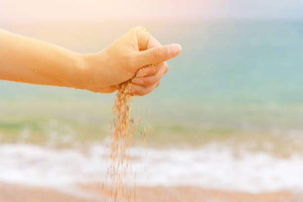 Close-up of a woman's hand releasing falling sand. Sand flows through your hand against the background of sea sand on the beach. The concept of a summer beach holiday