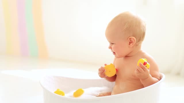 a little baby girl of six months with blue eyes bathes in a bubble bath and plays with rubber toys with ducks, smiles and rejoices splashing in the water, a happy baby washes