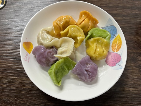 Stock photo showing an evening meal of steamed dumplings (Momos), filled with mixed vegetables and pork drizzled with chilli oil and garnished with chopped spring onion and coriander leaves on plate with red and orange spicy dipping sauce.