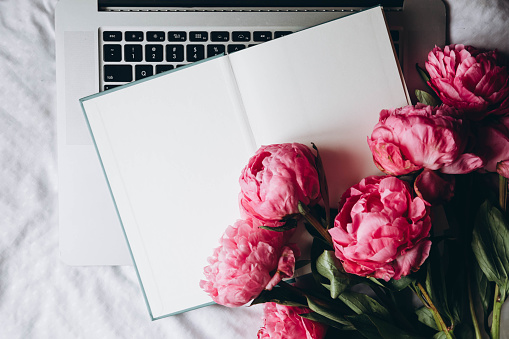 Opened notebook, laptop and pink peony flowers on the white bed sheet. Good morning and weekend concept in flat lay style, copy space.