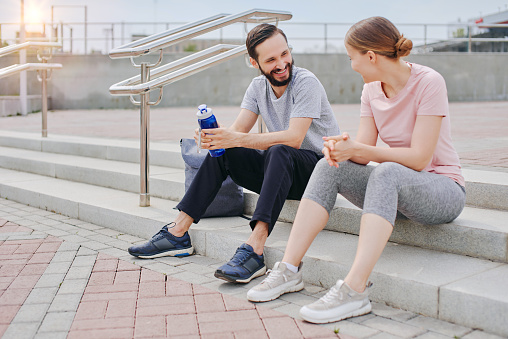 Happy man and woman in a simple sportswear preparing for outdoor jogging and work out, sitting on the stairs outside summertime
