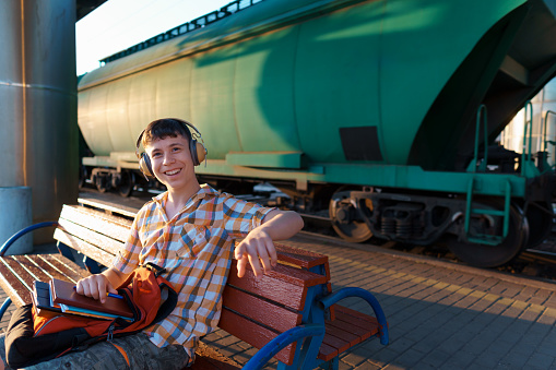 a student poses at a railway station, a boy waits for a train on the platform, listens to music with headphones, reads books and does homework, goes to study, the concept of education