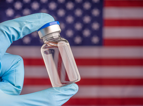 Hand in a blue glove holding a vaccine vial against the American flag background.  Close-up. Medical and vaccination concepts.