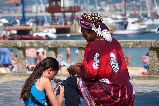 Baiona, Pontevedra, Spain; July 17, 2022; a woman of color braids braids on the street for a young white woman while she looks at her cell phone