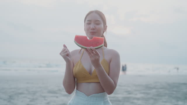 Young Asian Woman in Yellow Bikini Savoring Watermelon on a Beach at Sunset