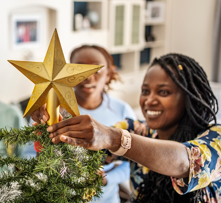 Happy african woman placing a golden star on the top of Christmas tree with her daughter standing by at home during holidays