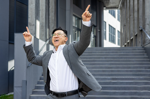 Joyful businessman celebrating success outside office building, pointing upwards with excitement and achievement in urban setting.