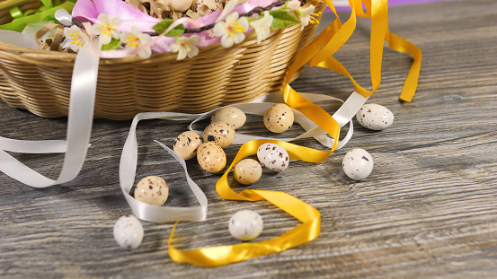 Small delicate pastel colored motley variegated spotted colored quail eggs in the wicker basket nest decorated with ribbons, spikelets and dried gypsophila flowers. Easter celebration concept. High quality 4k footage. Close up. Happy Easter. Holiday festive postcard bunny ears wicker basket paper letters gray wooden table.