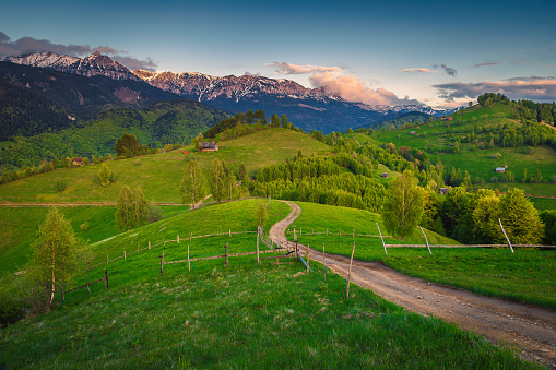 Spectacular curved rural road on the hills and majestic high mountains in background at sunset, Bucegi Mountains, Carpathians, Romania, Europe