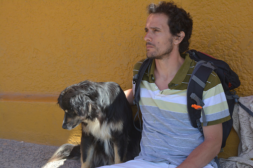 serious dark-haired young adult 30-40 years old with backpack sitting with a dog with yellow wall in the background