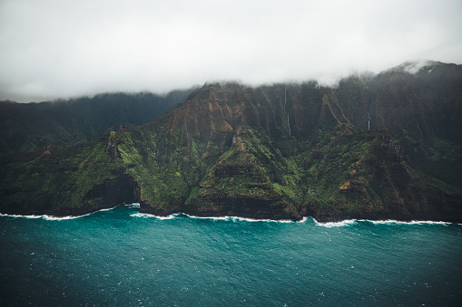 Experience the awe-inspiring grandeur of the Nā Pali Coast on the island of Kauai, Hawaii, with this breathtaking aerial image captured from a helicopter. The colossal volcanic cliffs, adorned with a kaleidoscope of vibrant hues including green, red, orange, and yellow, command attention and inspire wonder. Below, pristine sandy beaches beckon beneath the towering cliffs, offering a stark contrast to the rugged terrain above. Atop the volcano mountains, clouds dance and mingle, casting shadows and adding a dramatic touch to the scene. The azure blue waters of the ocean complete the picture-perfect vista, enhancing the timeless beauty of Kauai's iconic coastline. Immerse yourself in the splendor of Nā Pali Coast, where nature's magnificence unfolds in every breathtaking detail.