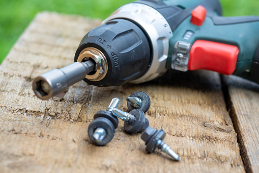 A close-up of an electric screwdriver with a nozzle on lies on a wooden background. Next to it, roofing screws on metal are laid out in a row. The concept of repair and construction