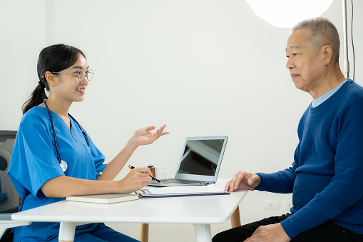 Asian professional female doctor talking with male patient about his pain and symptoms while giving consultation at table in examination room at health care hospital