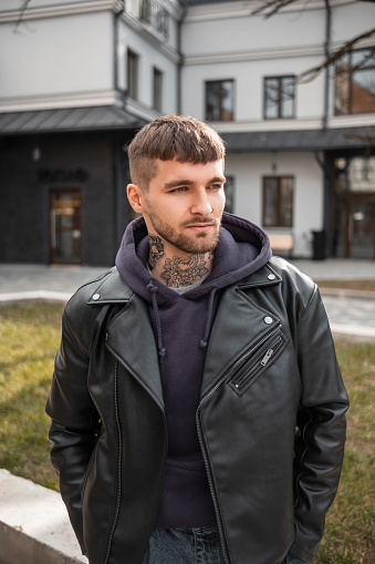 Beauty portrait of a handsome stylish casual hipster man with hair, beard, and neck tattoo in a trendy black leather jacket and hoodie walking down the street