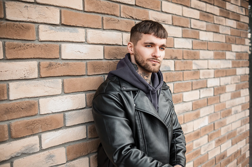 Stylish handsome young hipster man with hair and tattoos on his neck in a fashionable black leather jacket and hoodie standing near a brick wall