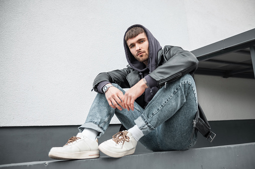 Fashionable handsome young hipster man wearing trendy clothes with jacket, hoodie, jeans and sneakers sitting near the white wall on the street