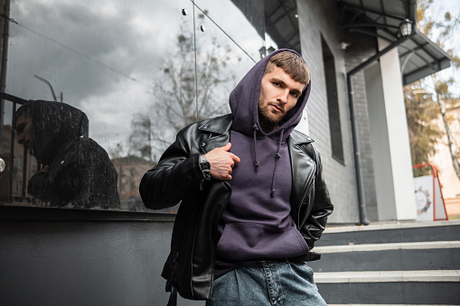Stylish handsome young male hipster with a fashion purple hoodie mockup wears a black rock leather jacket near a modern black building on the street