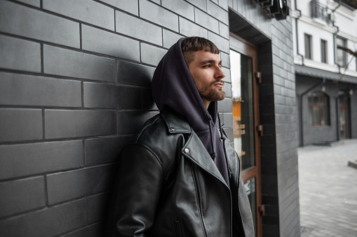 Stylish men's street portrait of a handsome brutal man in fashionable clothes with a black rock leather jacket and hoodie stands near a black brick modern building