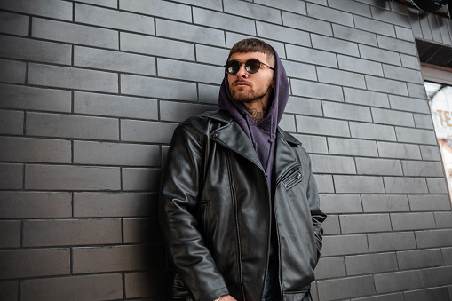 fashionable handsome hipster man with fashionable round sunglasses wearing fashionable leather jacket and hoodie stands near a black brick wall on the street