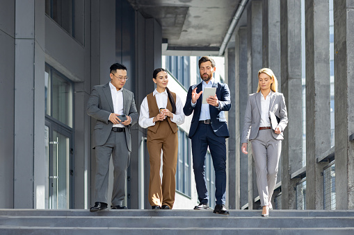 A group of four business professionals confidently strolling down steps outside a modern office building, engrossed in discussion with digital tablets and smartphones in hand.