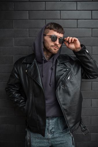 Fashion street portrait of handsome man hipster with cool stylish sunglasses in trendy leather jacket with hoodie poses near a black modern brick background