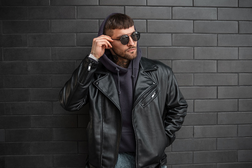 Cool trendy hipster model man in fashionable clothes with leather jacket and hoodie adjusts his stylish sunglasses near a black brick wall