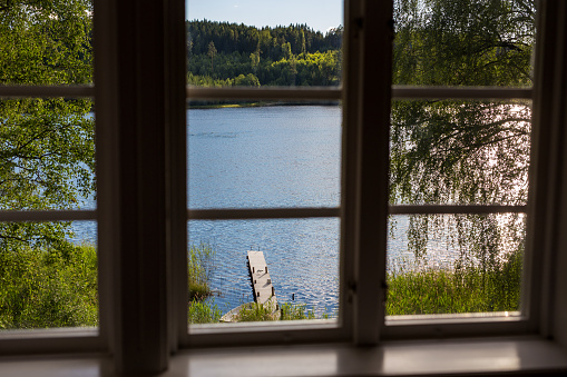 View from inside through a window during a summer day of a deserted Swedish lake with wooden jetty and rowboat and forest in the background