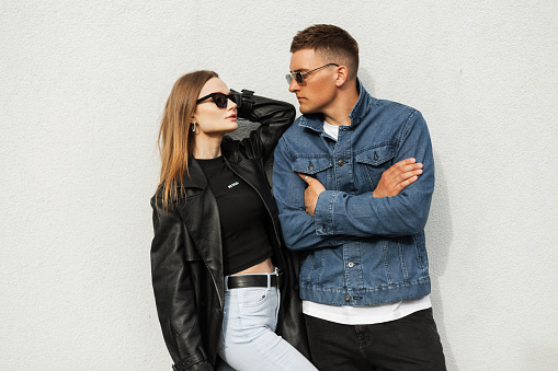 Fashionable couple beautiful model woman and handsome stylish man with vintage sunglasses in casual urban clothes with leather black coat and denim jacket poses near the wall on the street