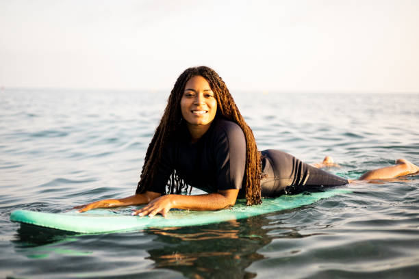 istock A young dark-skinned woman posing on a surfboard in the sea dressed in a wetsuit while looking at the camera happily. African women with long hair surfing. Water sports. Calm sea to practice. 2109442871