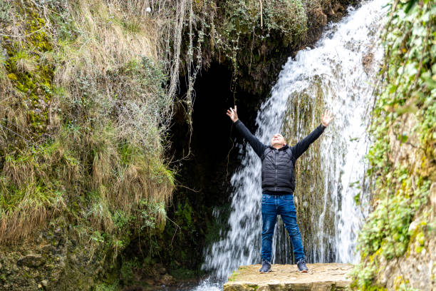A middle-aged man is euphoric with raised arms near a waterfall in a rural setting in the middle of nature, Tobero Burgos - Spain