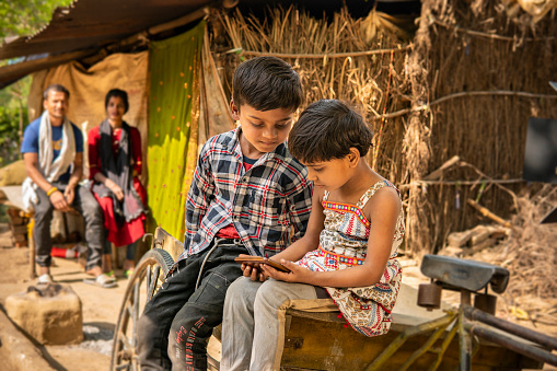 Outdoor portrait of two happy rural Indian children sitting together on old rickshaw cart outside their house and using smartphone in leisure time. Their parents sitting behind and looking at them.