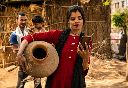 Portrait of happy traditional, rural indian married young woman carrying drinking water clay pot while using smartphone at day time and her husband stands behind with child in the village.