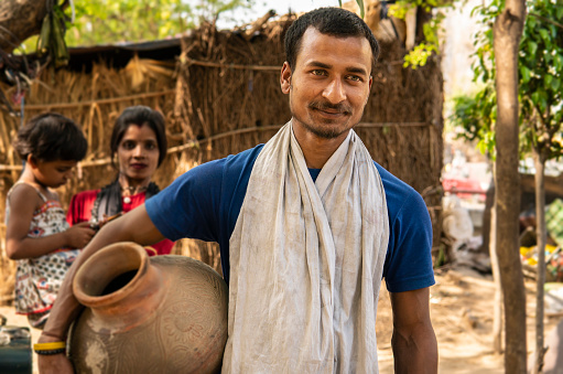Portrait of happy traditional, rural indian young man carrying drinking water clay pot and contemplating while looking away and her wife stands behind with child in the village.