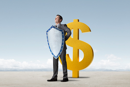 A man holding a medieval shield stands confidently and protectively in front of a tall gold dollar sign.