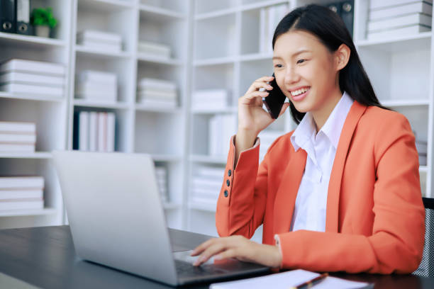 istock A woman is talking on her cell phone while sitting at a desk with a laptop 2109592582