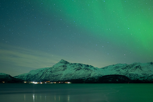 Green Northern Lights Dancing in Arctic skies above a Norweigan lake and mountain.  Beautiful and tranquil Aurora Borealis in the arctic circle in Northern Norway with snowcapped mountains in the background. Shot on Sony mirrorless camera in the late fall near Tromso.