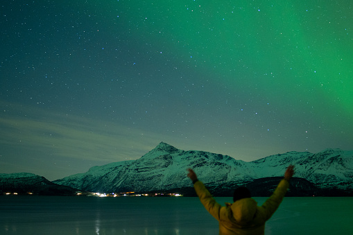 Person reaching up to green Northern Lights Dancing in Arctic skies above a Norweigan lake and mountain.  Beautiful and tranquil Aurora Borealis in the arctic circle in Northern Norway with snowcapped mountains in the background. Shot on Sony mirrorless camera in the late fall near Tromso.  Man in yellow jacket looking up at the sky at northern lights.