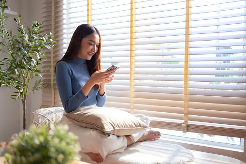 Smiling Asian cheerful adult female enjoy using smartphone surf internet social media relax casual daily technology in life,woman use smartphone weekend on wooden bench near window in living room at home